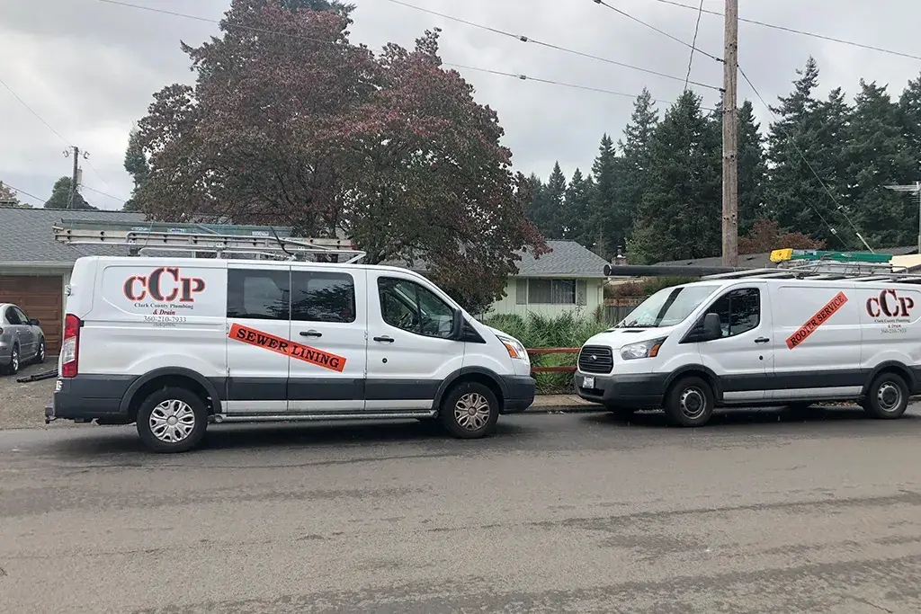 Two white Clark County Plumbing & Drain service vans parked on a residential street, featuring "Sewer Lining" and "Rooter Service" decals.