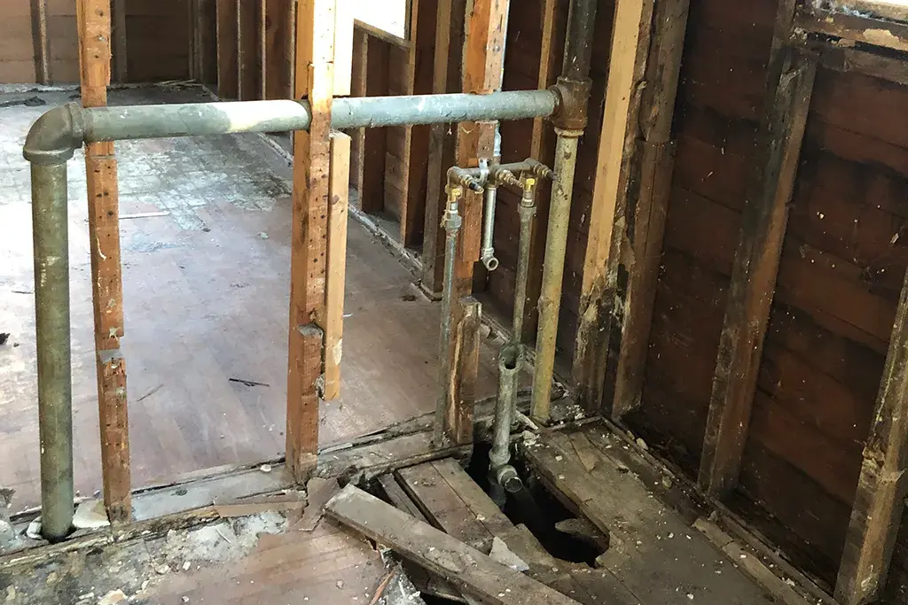 Interior view of a home renovation showing old galvanized water lines and drain pipes exposed within the wooden wall framing.