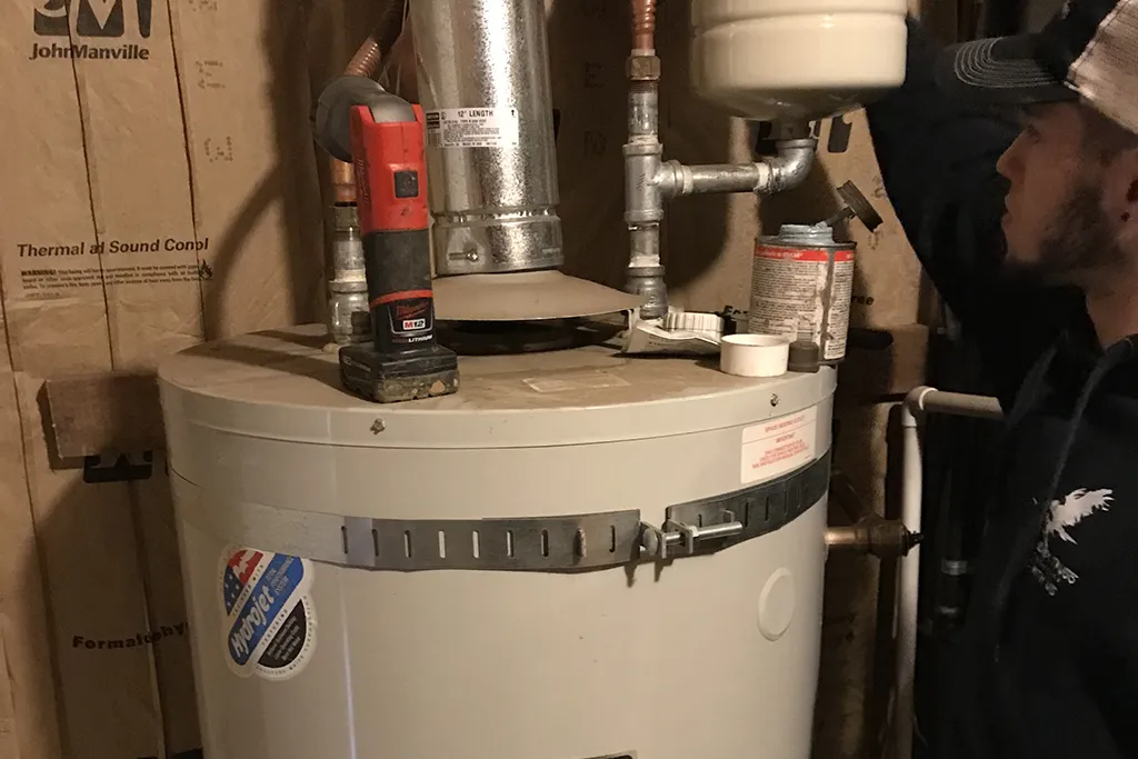 A technician performing maintenance on a residential water heater, with tools and sealant staged on top of the tank.