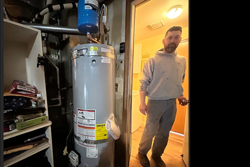 A professional plumber standing next to a newly installed tall cylindrical water heater in a residential basement.