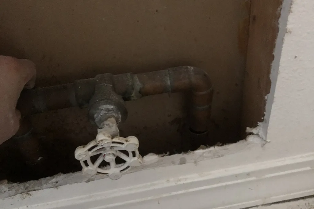 Close-up of an old copper water main shut-off valve with a white circular handle located inside a wall cutout.