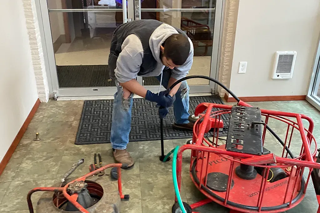 A technician using a red Maxliner drain cleaning machine inside a commercial or residential lobby to clear a blockage.