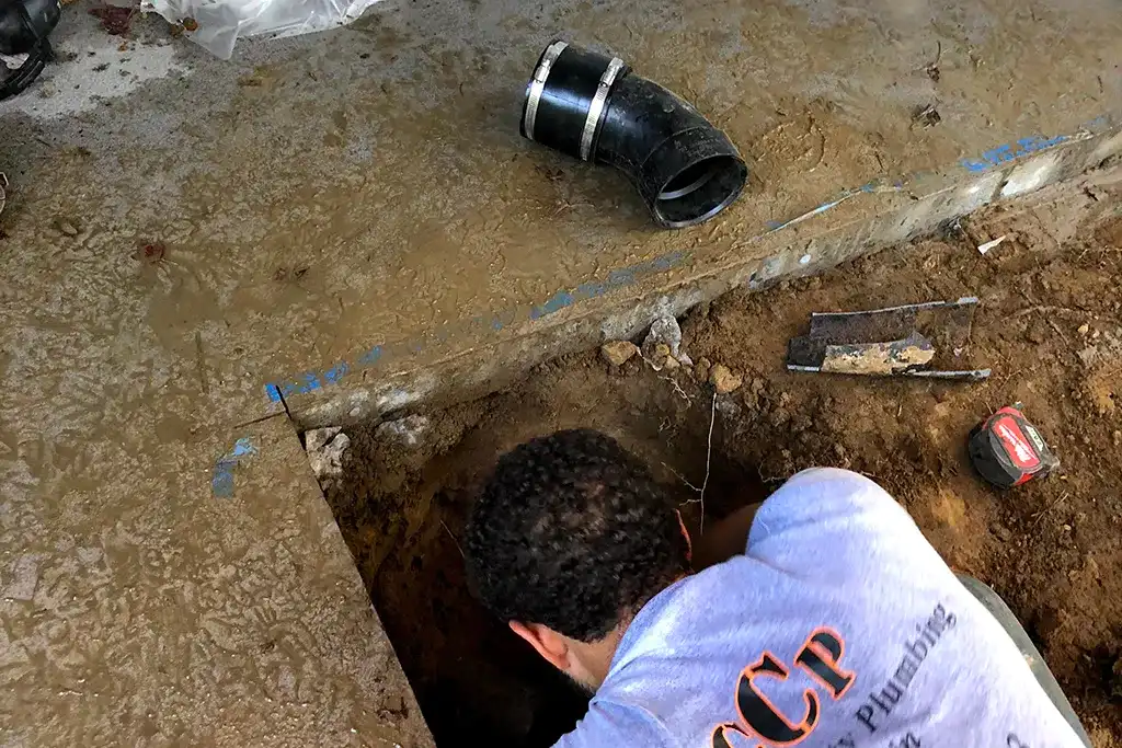 A technician working in a small square cutout in a concrete floor to access a pipe for repair.