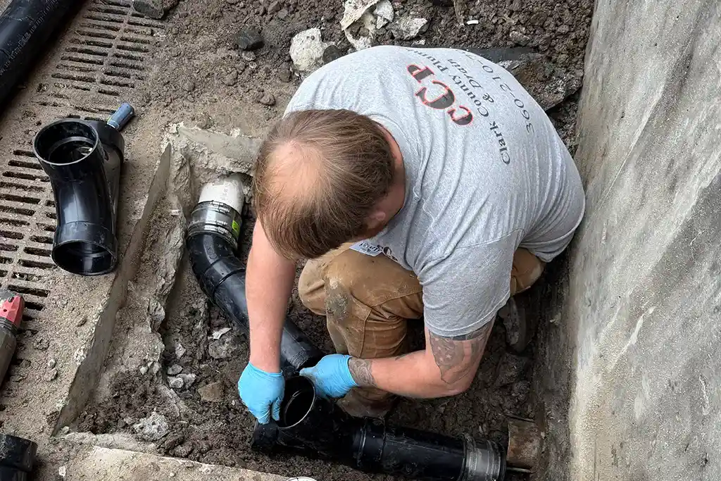 A technician in blue gloves installing a new black ABS sewer cleanout and pipe fittings in a basement excavation.